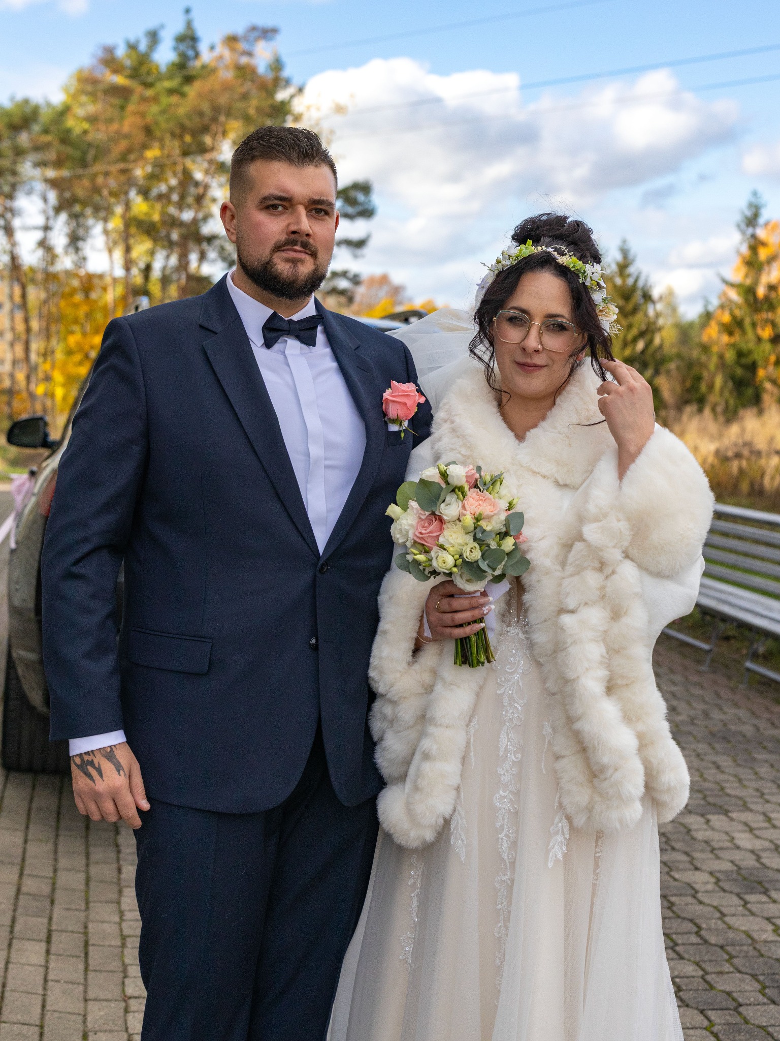 Para młoda w eleganckich strojach ślubnych pozuje na zewnątrz, trzymając bukiet. W tle widoczne drzewa i fragment samochodu.