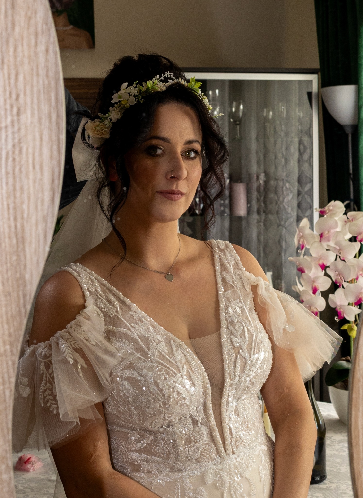Panna młoda w koronie z kwiatów i sukni ślubnej z koronkowymi zdobieniami, odbijająca się w lustrze. W tle storczyki i elementy wystroju wnętrza.