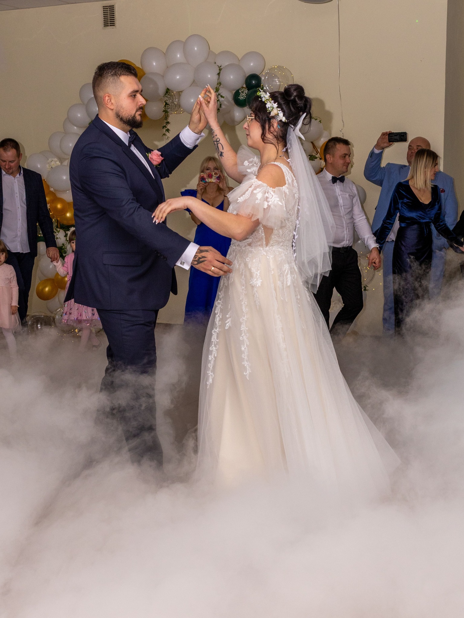 Pierwszy taniec pary młodej w obłokach dymu, goście obserwują w tle, dekoracje balonowe, Piotrków Trybunalski. Panna młoda w sukni z koronką, pan młody w granatowym garniturze.