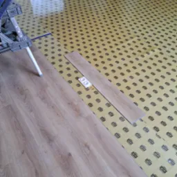 Proces układania paneli podłogowych: widoczny fragment ułożonej podłogi, podkład z nadrukowanymi liniami pomocniczymi oraz dwa panele i akcesoria do montażu