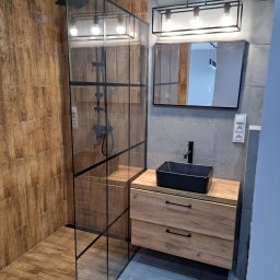 Usługi Remontowo-Budowlane Paweł Majczyna - Nowoczesna łazienka z kabiną prysznicową typu walk-in z czarnymi profilami, umywalką nablatową w kolorze czarnym i szafką z drewnopodobnym frontem oraz szarą płytką na ścianach.