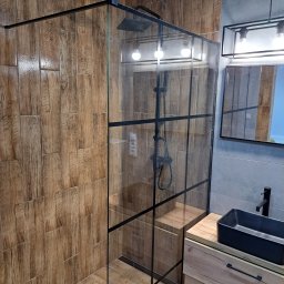 Usługi Remontowo-Budowlane Paweł Majczyna - Nowoczesna kabina prysznicowa typu walk-in z czarnymi profilami, drewnopodobne płytki na ścianach i podłodze, czarna umywalka nablatowa na szafce z drewnianym frontem, szara ściana w tle.