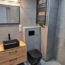 Nowoczesna łazienka w szarościach z drewnianą szafką pod umywalkę, czarną armaturą i grzejnikiem drabinkowym.