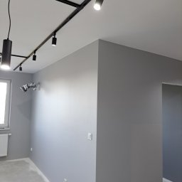 Usługi Remontowo-Budowlane Paweł Majczyna - Wnętrze pokoju po malowaniu ścian na szaro, z zamontowanym czarnym systemem szynoprzewodów oświetleniowych na suficie, widoczne gniazdka elektryczne na ścianie i zabezpieczona folią klimatyzacja.