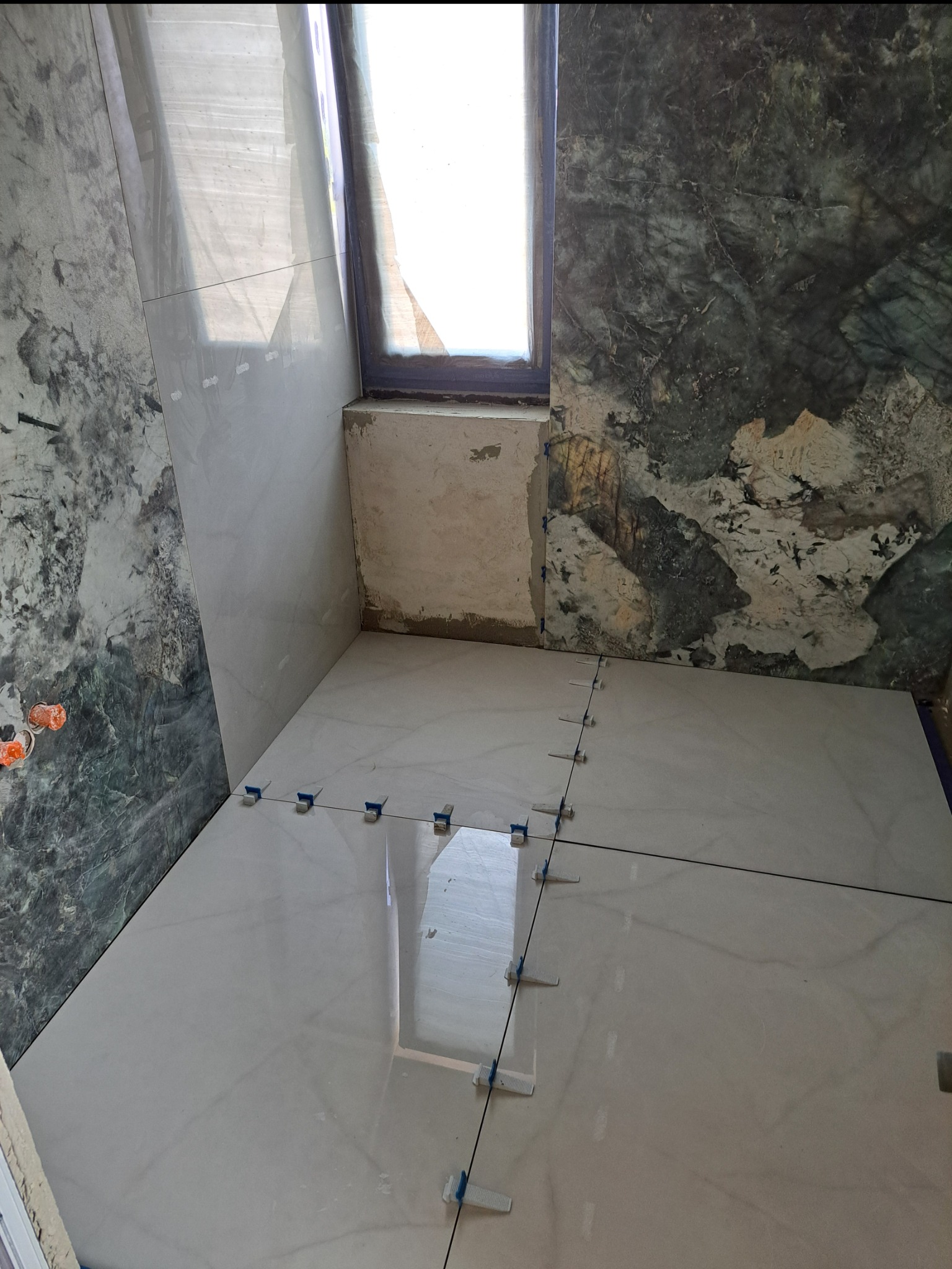 Błyszczące, kremowe płytki podłogowe w trakcie układania, z systemem poziomowania płytek, na tle ściany z zielonego marmuru i surowego muru pod oknem.