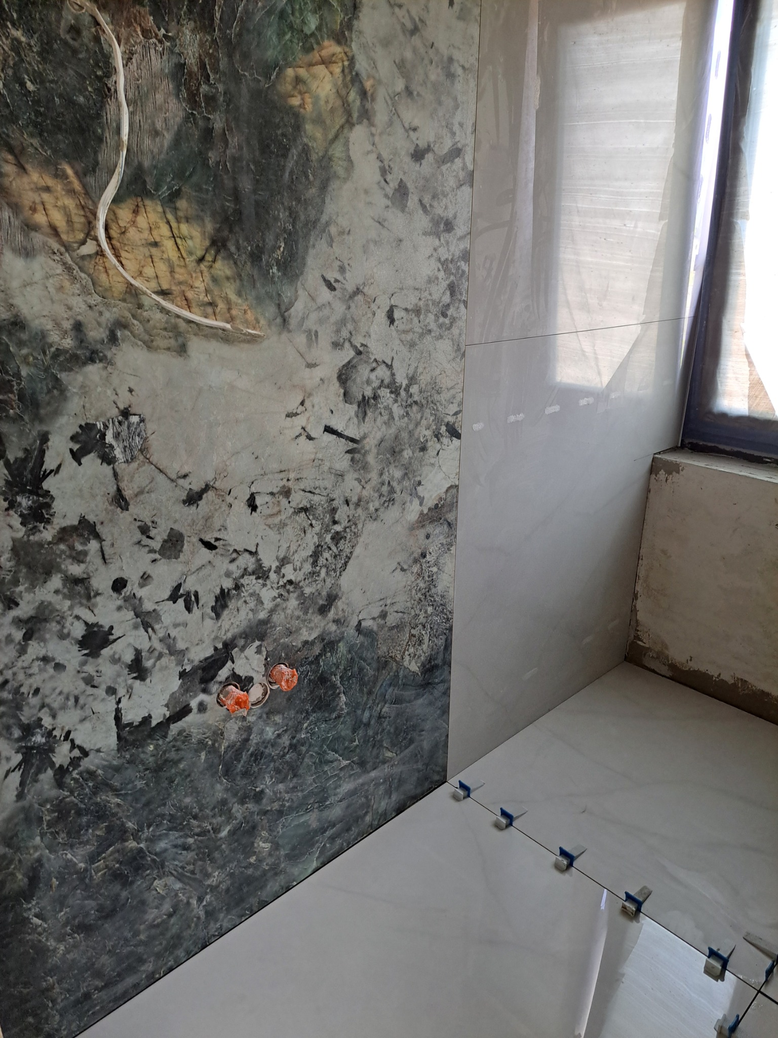 Ściana z częściowo ułożonymi płytkami o wzorze marmuru, widoczne puszki elektryczne i poziomujące klipsy na podłodze.