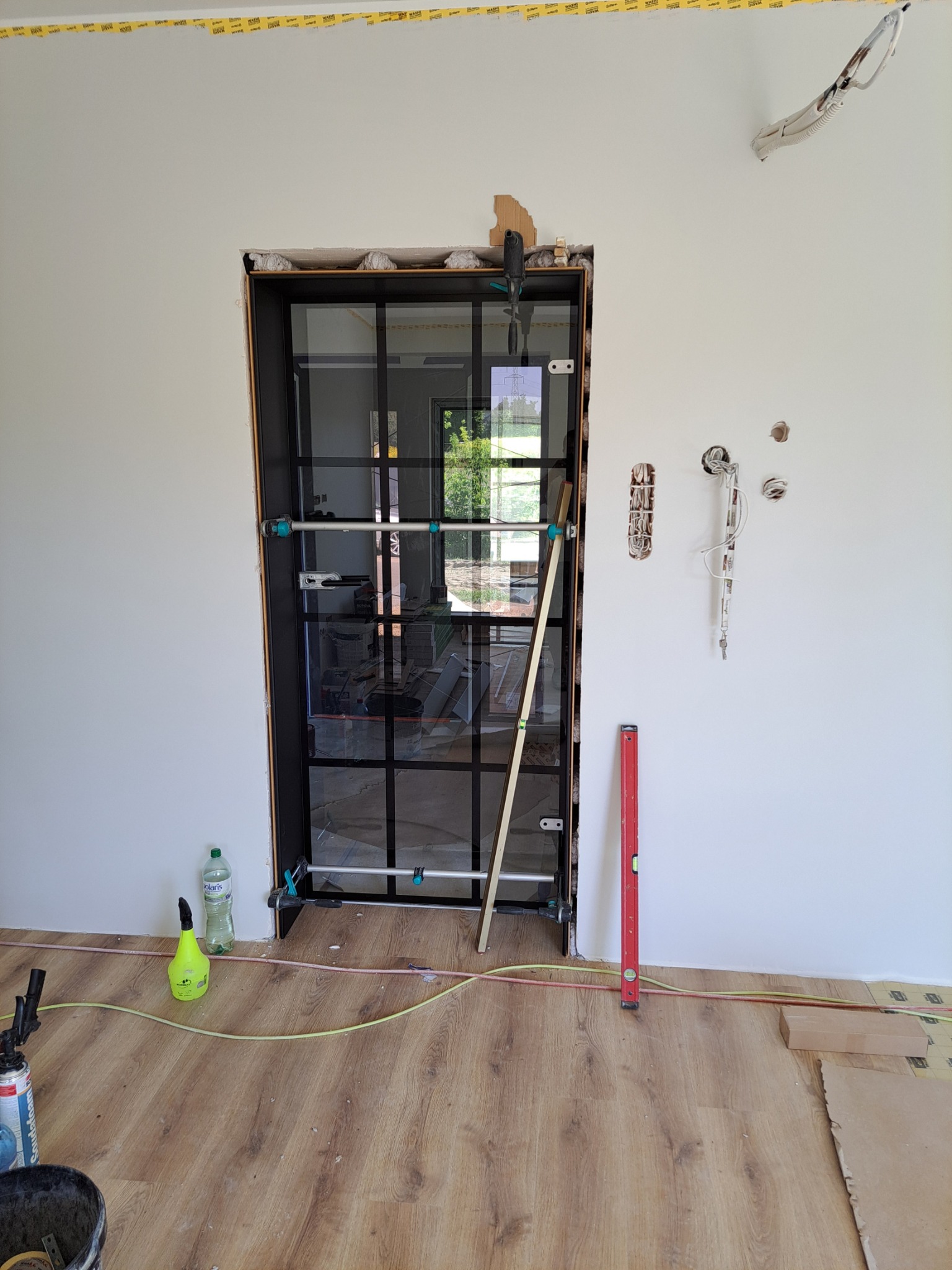 Montaż czarnej, przeszklonej ścianki działowej w surowym otworze, widoczne narzędzia, poziomica, butelka z płynem, niezabezpieczone przewody elektryczne w ścianie, fragment ułożonej podłogi panelowej.