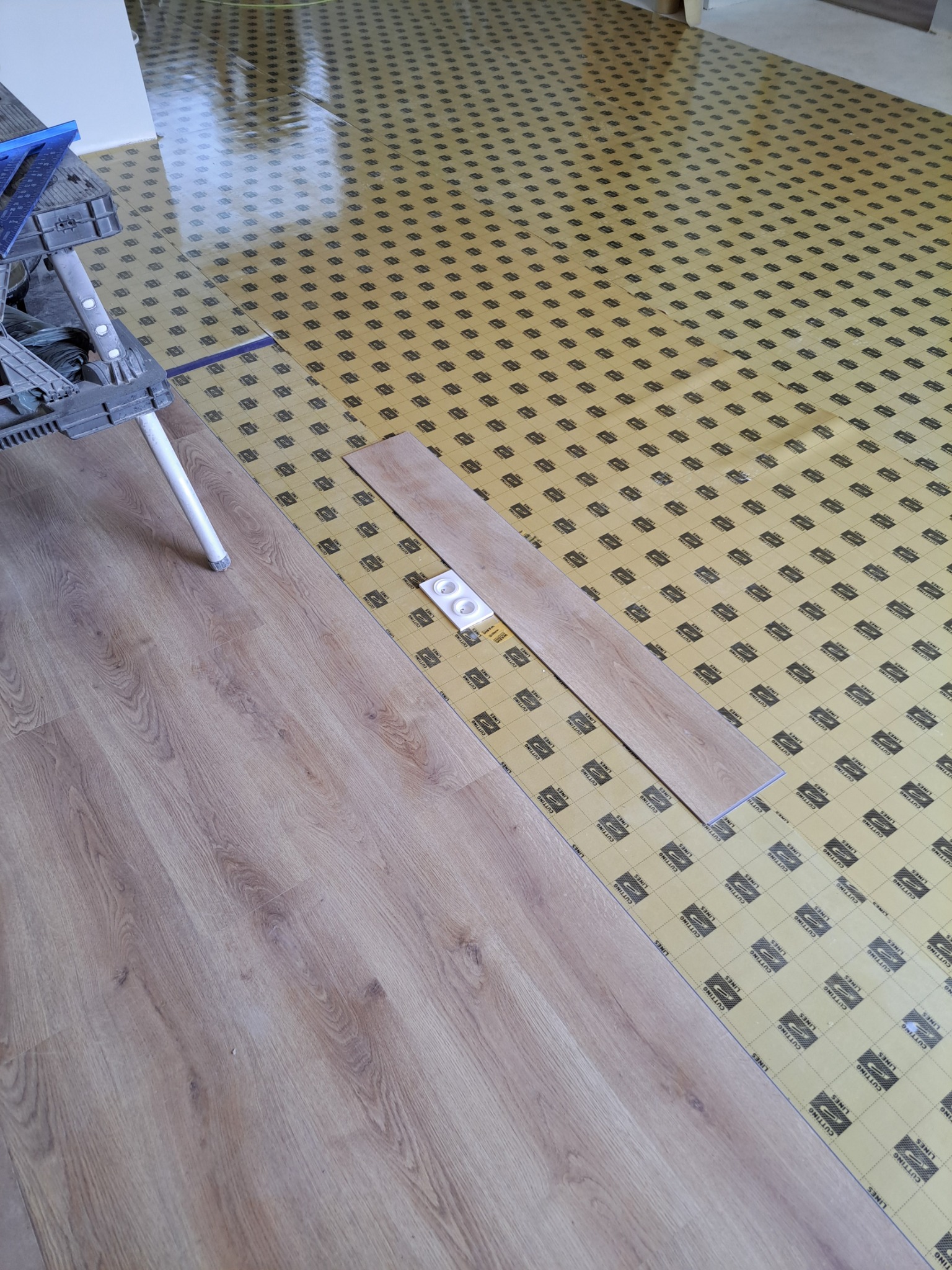 Proces układania paneli podłogowych: widoczny fragment ułożonej podłogi, podkład z nadrukowanymi liniami pomocniczymi oraz dwa panele i akcesoria do montażu