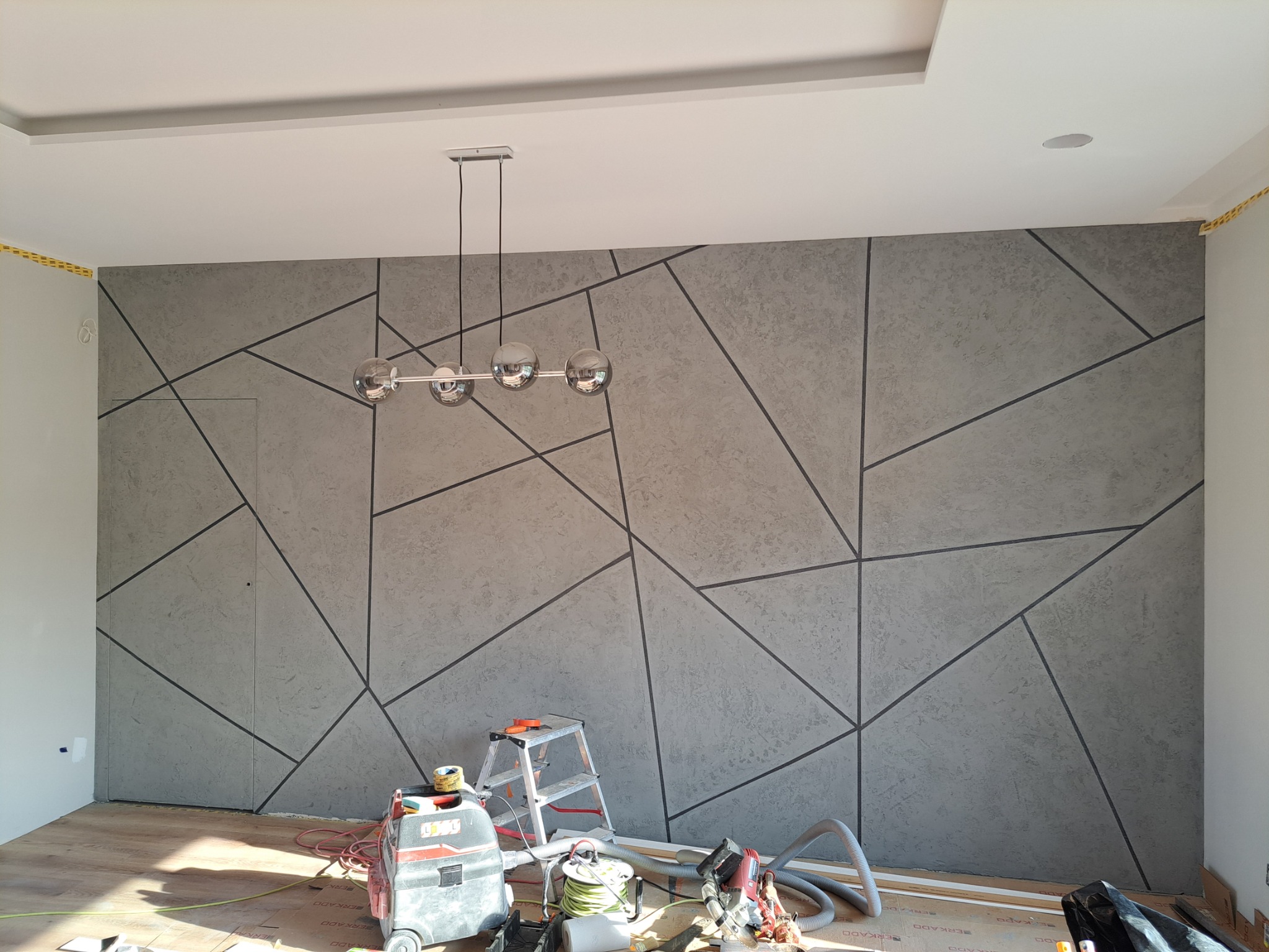 Ściana z szarą dekoracyjną strukturą i geometrycznym wzorem, podświetlona wiszącą lampą z czterema szklanymi kulami, narzędzia i drabina na podłodze zabezpieczonej kartonem.