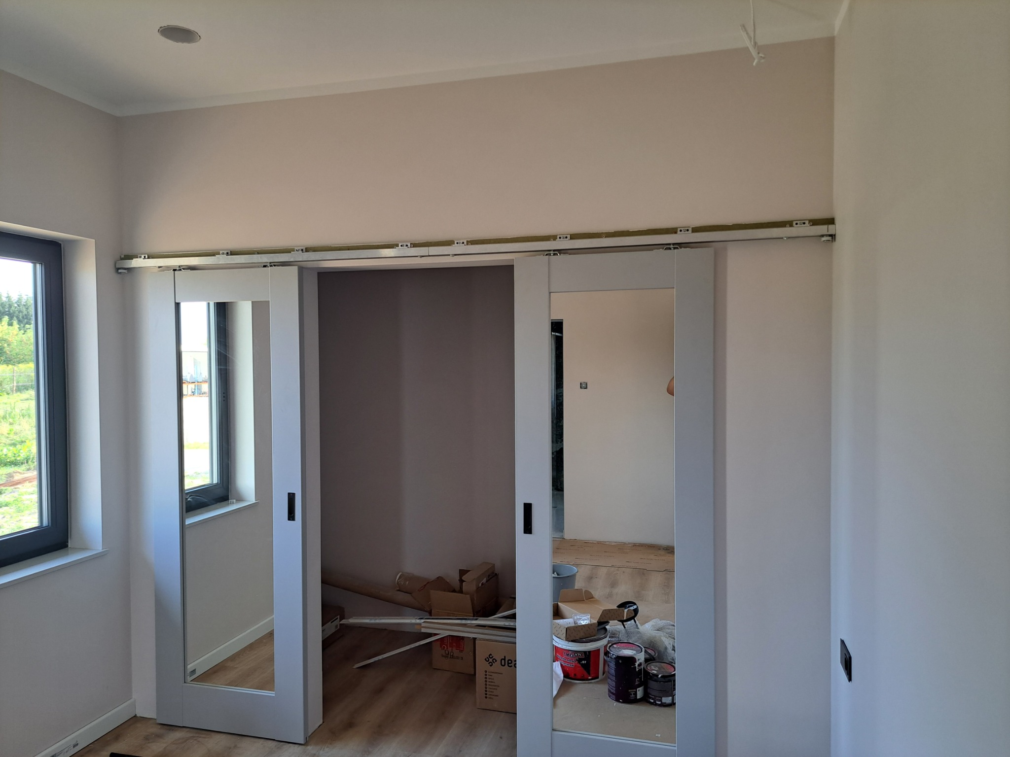 Montaż przesuwnych drzwi z lustrami w jasnym pokoju, widoczne puszki z farbą i kartony na podłodze.