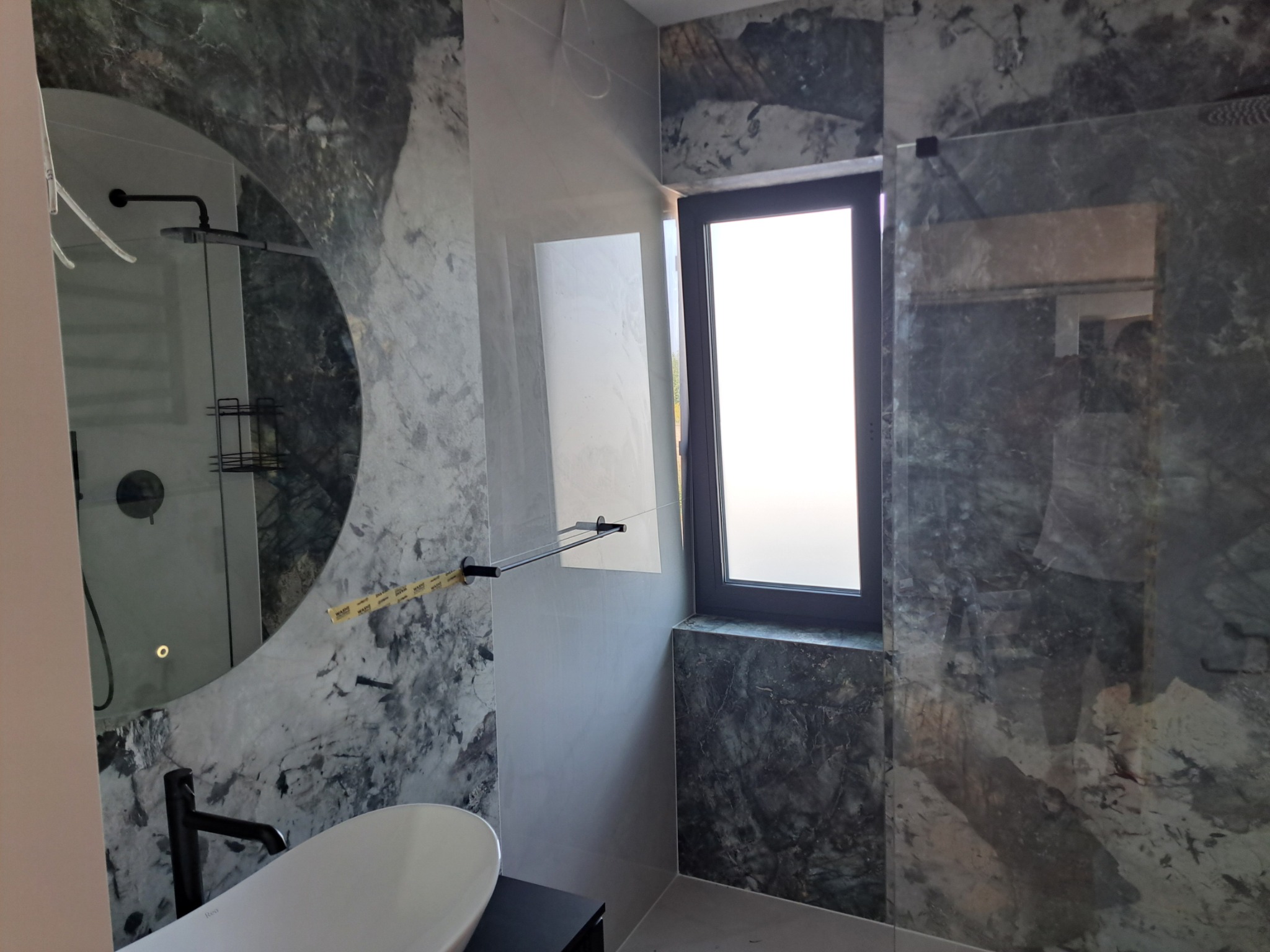 Nowoczesna łazienka z umywalką nablatową, okrągłym lustrem i ścianami wykończonymi płytkami imitującymi marmur.