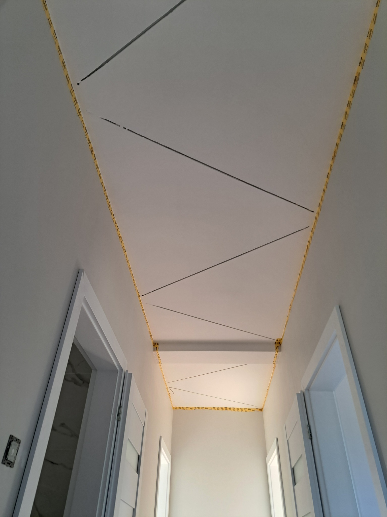 Przygotowanie sufitu do prac wykończeniowych: wyznaczone linie i zabezpieczające taśmy malarskie wzdłuż krawędzi, widoczne białe ściany i ościeżnice drzwiowe.