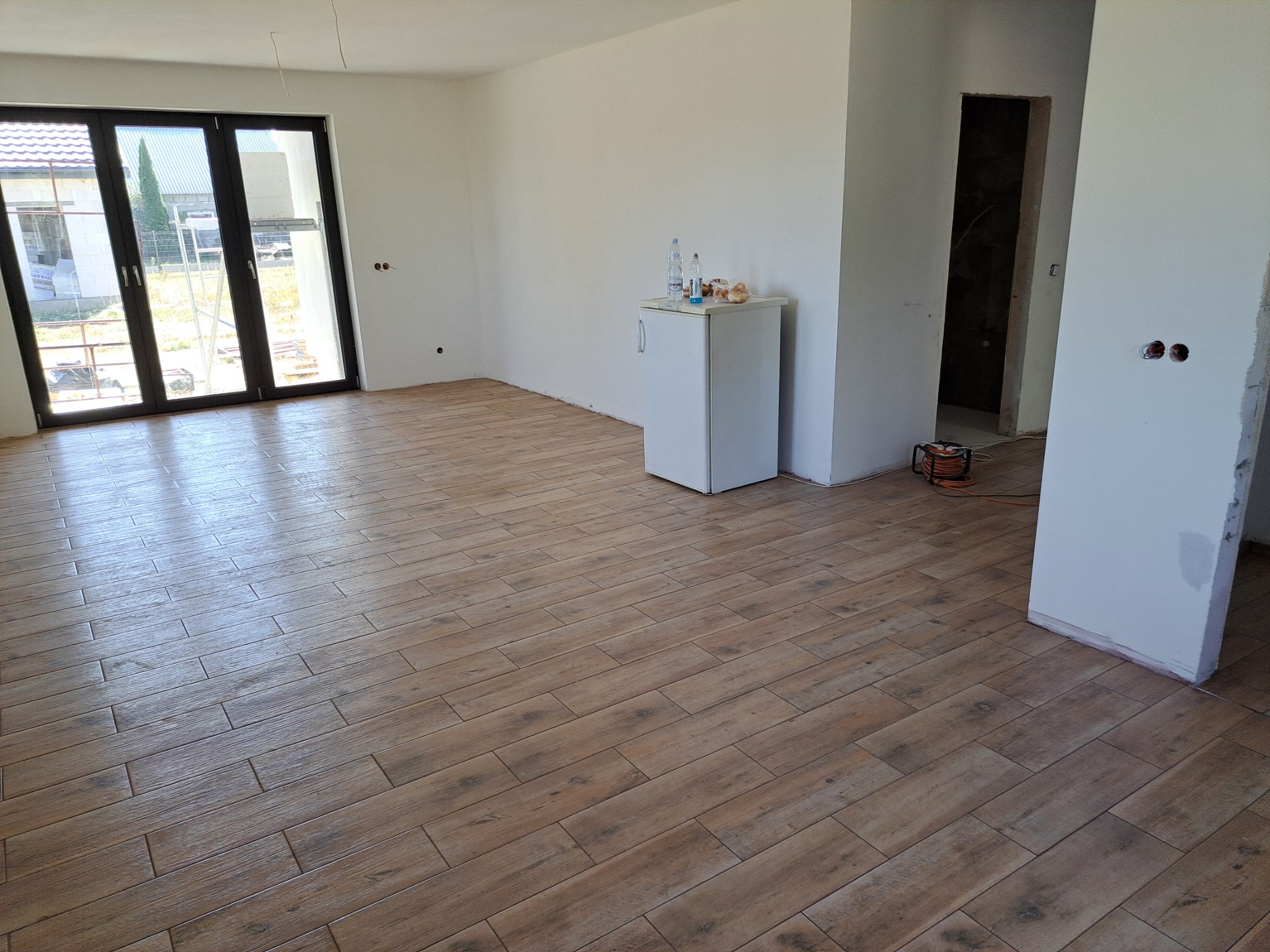 Salon po remoncie z brązowymi płytkami imitującymi drewno na podłodze, widoczne białe ściany, okno balkonowe i mała lodówka z napojami.
