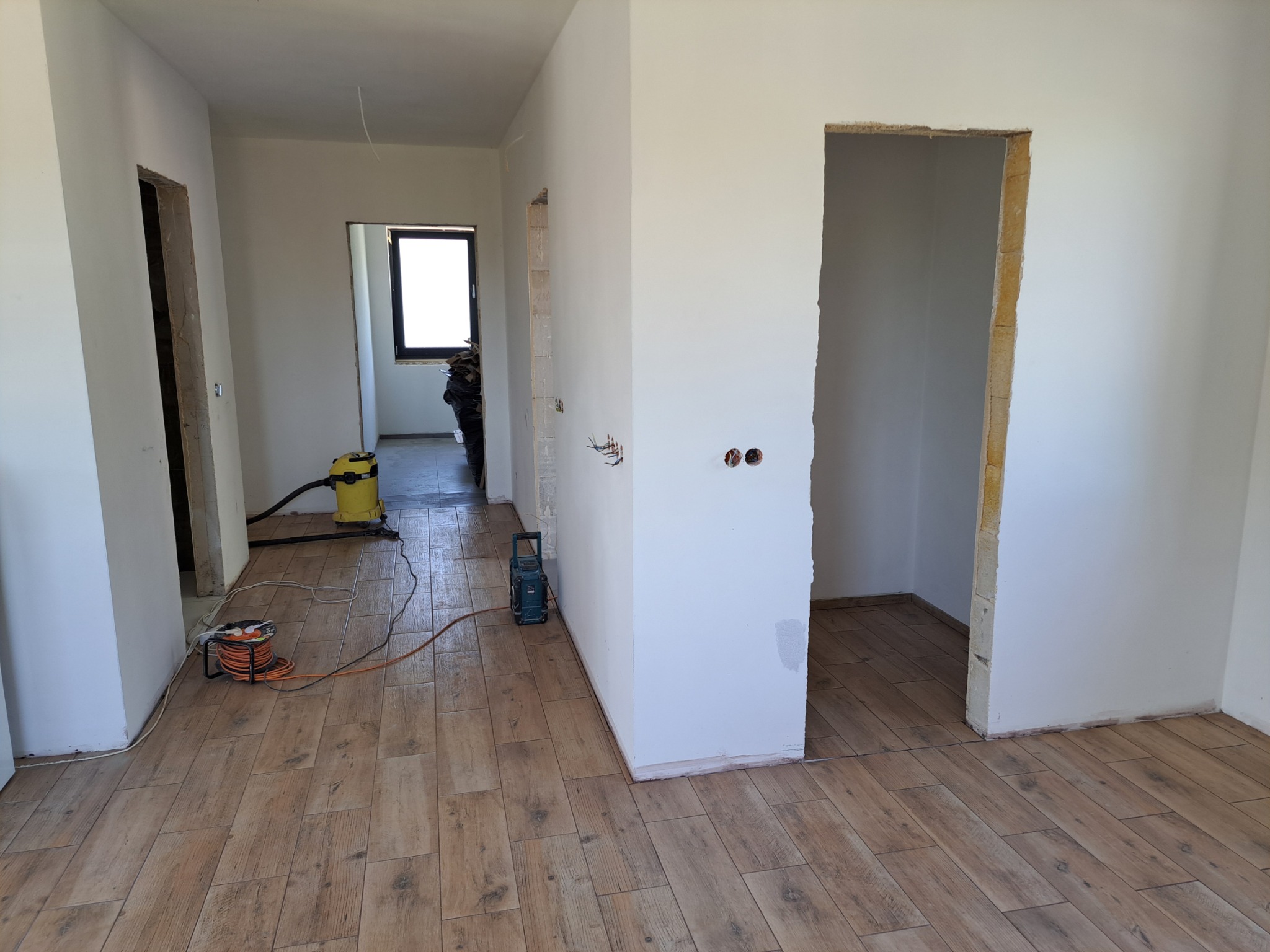 Wnętrze w trakcie prac wykończeniowych: ułożona podłoga z paneli imitujących drewno, świeżo pomalowane ściany, widoczne kable elektryczne wystające ze ściany, odkurzacz przemysłowy i zwinięty...