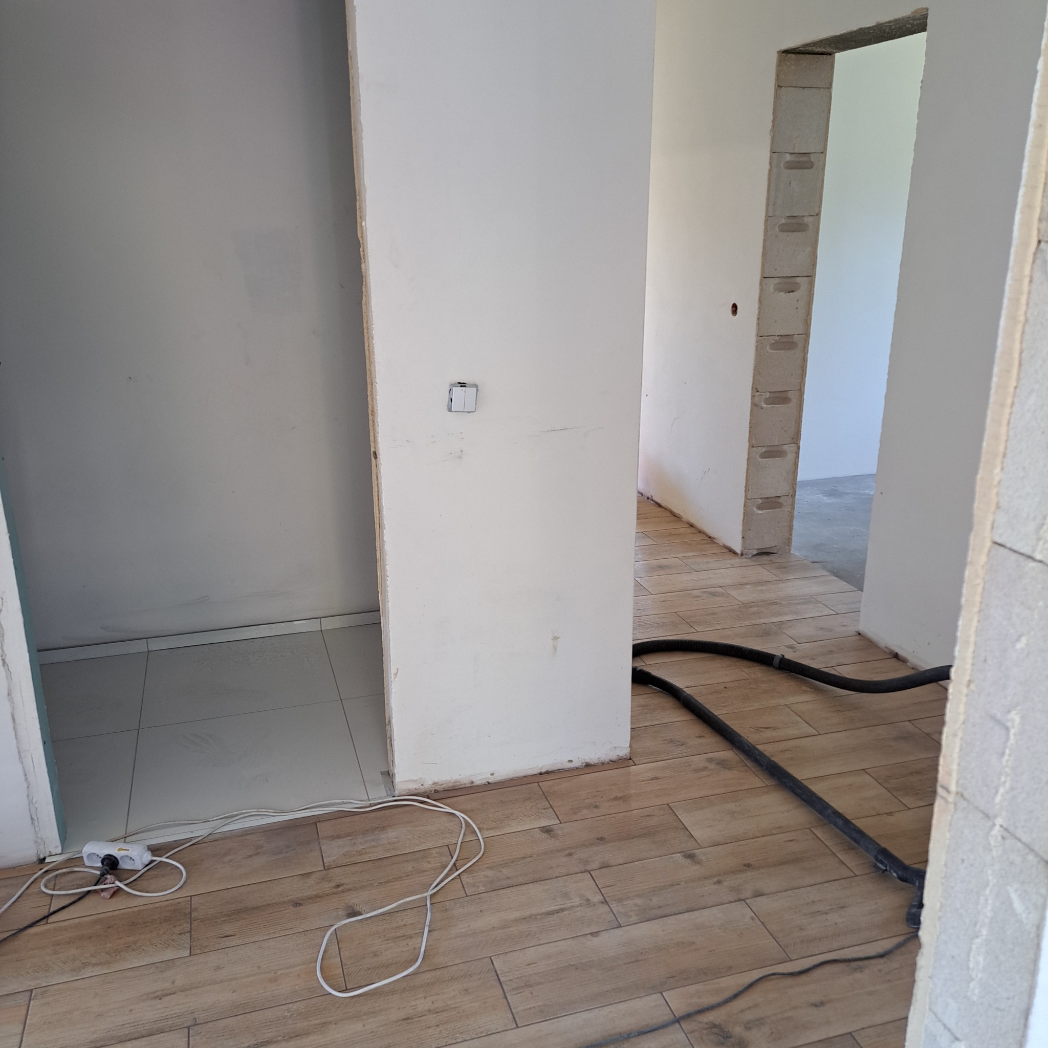Wnętrze w trakcie prac wykończeniowych: położona podłoga z paneli imitujących drewno, surowe ściany, widoczne przewody elektryczne i wąż od odkurzacza.