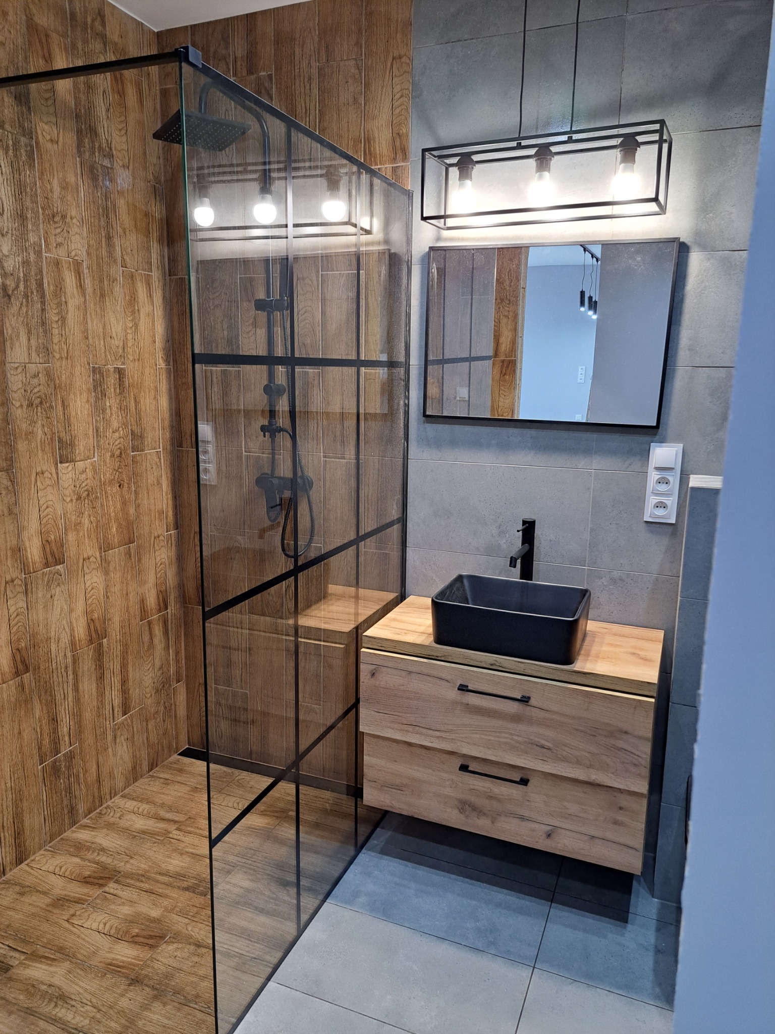 Nowoczesna łazienka z kabiną prysznicową typu walk-in z czarnymi profilami, umywalką nablatową w kolorze czarnym i szafką z drewnopodobnym frontem oraz szarą płytką na ścianach.
