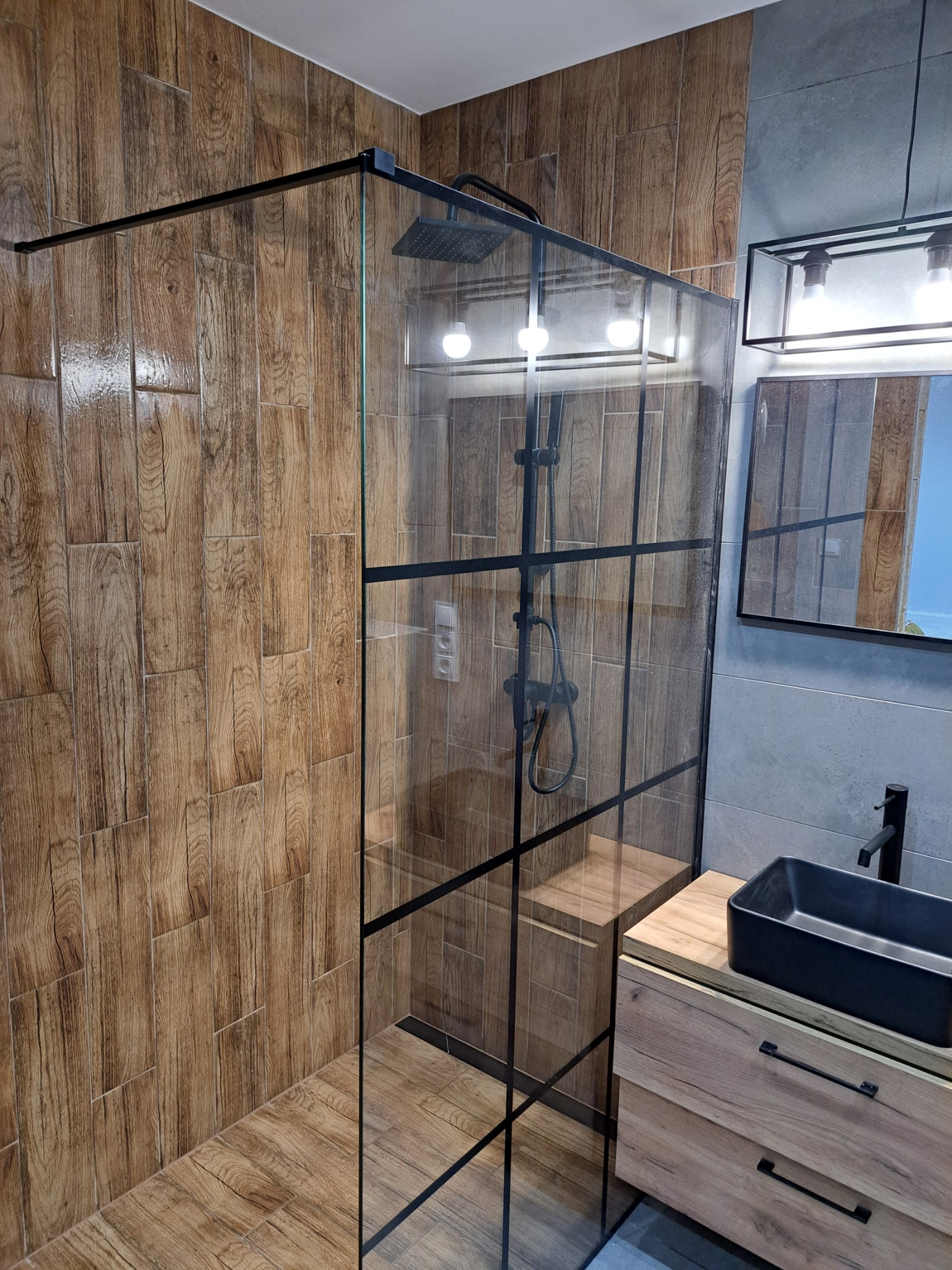 Nowoczesna kabina prysznicowa typu walk-in z czarnymi profilami, drewnopodobne płytki na ścianach i podłodze, czarna umywalka nablatowa na szafce z drewnianym frontem, szara ściana w tle.