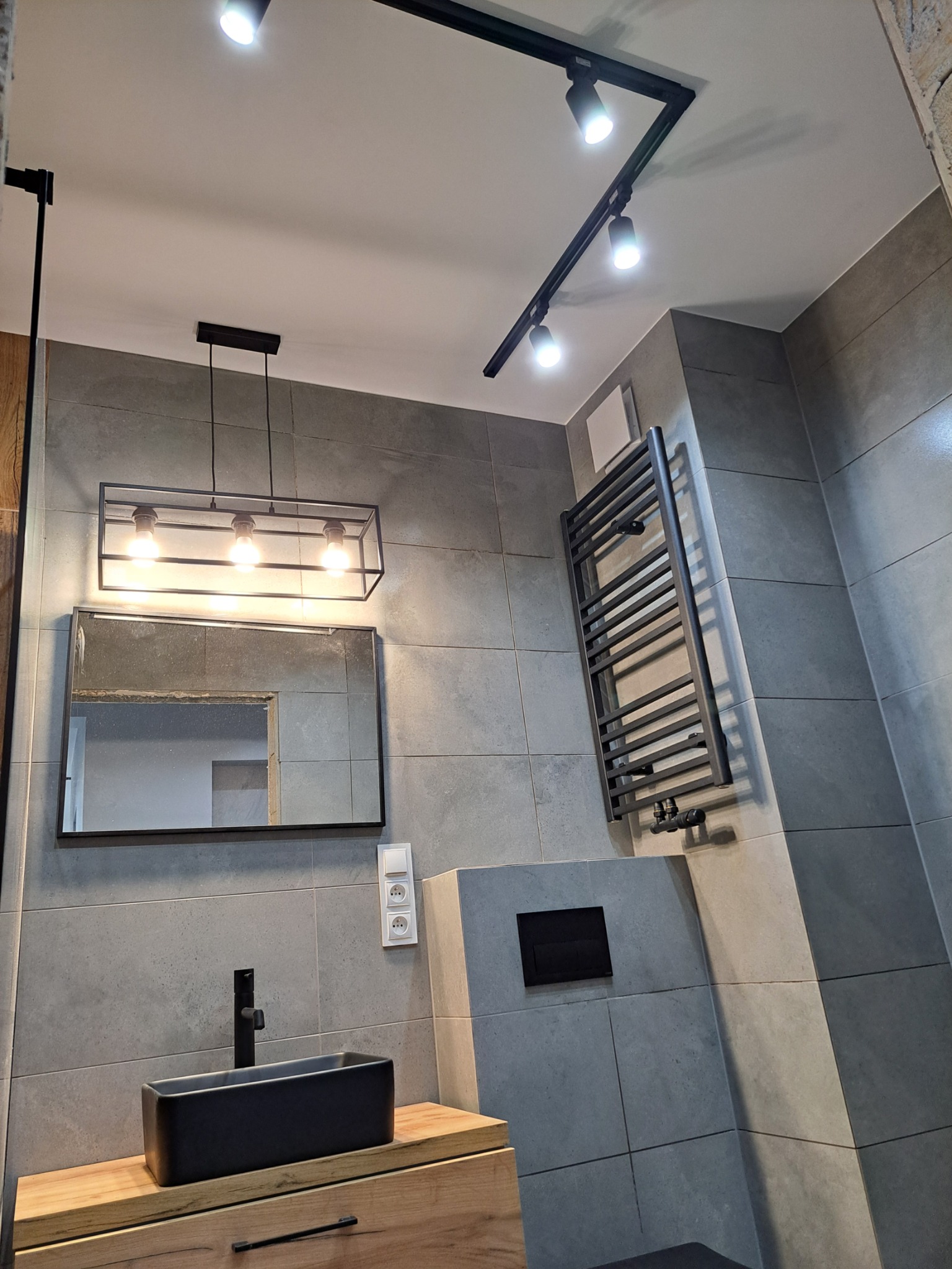 Nowoczesna łazienka z szarymi płytkami, czarną umywalką nablatową, grzejnikiem drabinkowym i oświetleniem punktowym na suficie.