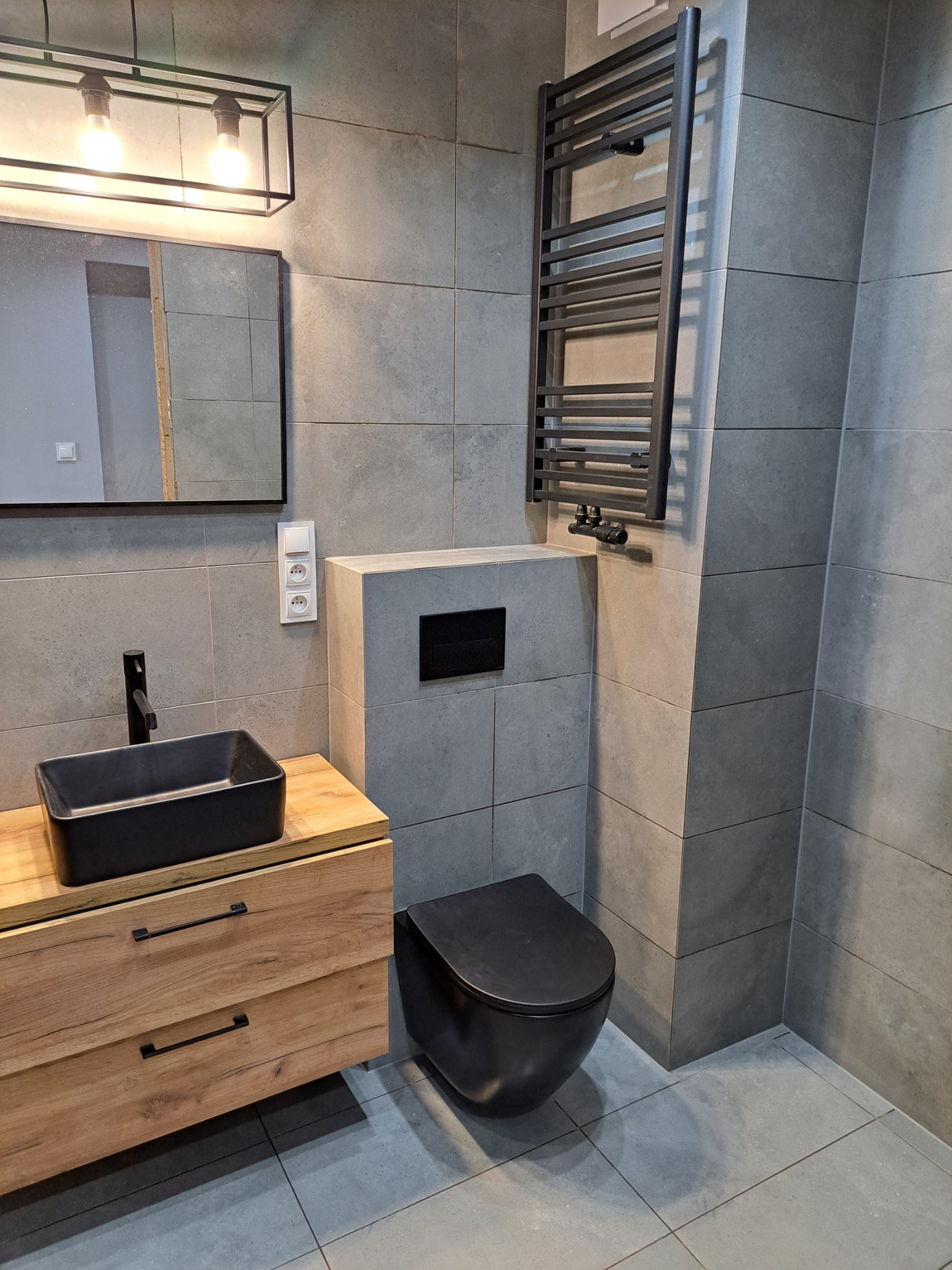 Nowoczesna łazienka w szarościach z drewnianą szafką pod umywalkę, czarną armaturą i grzejnikiem drabinkowym.