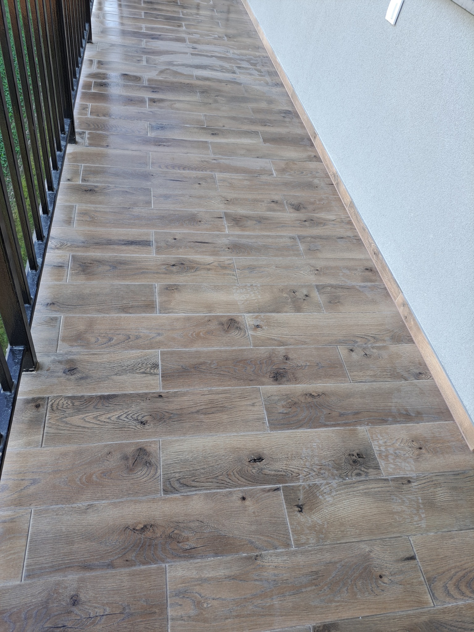 Wyłożony płytkami imitującymi drewno balkon z czarną, metalową barierką i jasną ścianą z listwą przypodłogową.