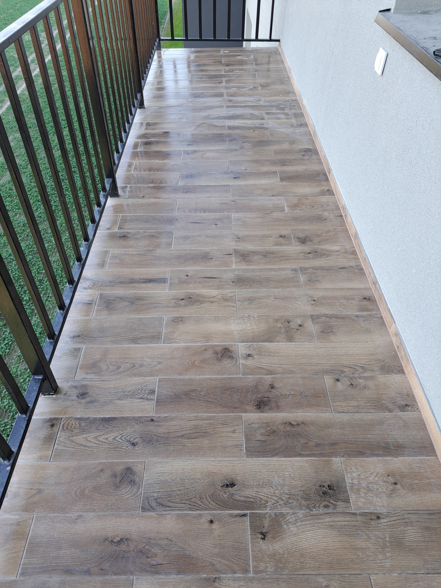 Długi balkon wyłożony płytkami imitującymi drewno, z metalową balustradą i widokiem na zieleń