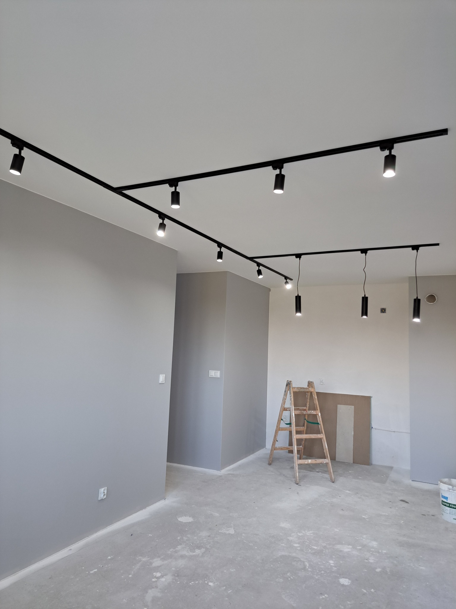 Szare ściany w trakcie prac wykończeniowych, widoczne czarne reflektory na szynoprzewodzie, drabina malarska i wiadro z gładzią.