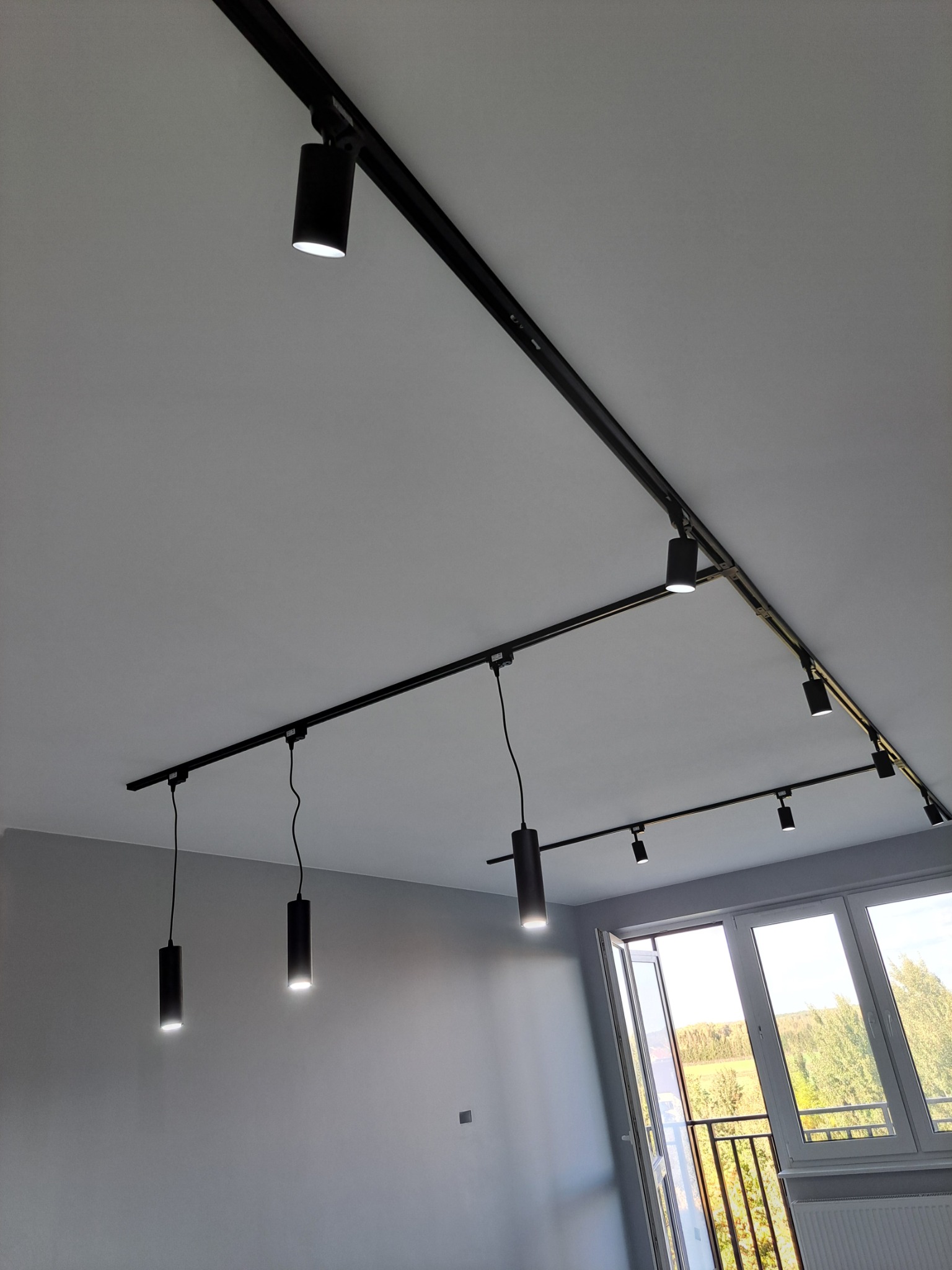 Nowoczesny system oświetlenia szynowego z czarnymi reflektorami cylindrycznymi, zamontowany na suficie w szarym pomieszczeniu z oknem balkonowym i widokiem na zieleń.