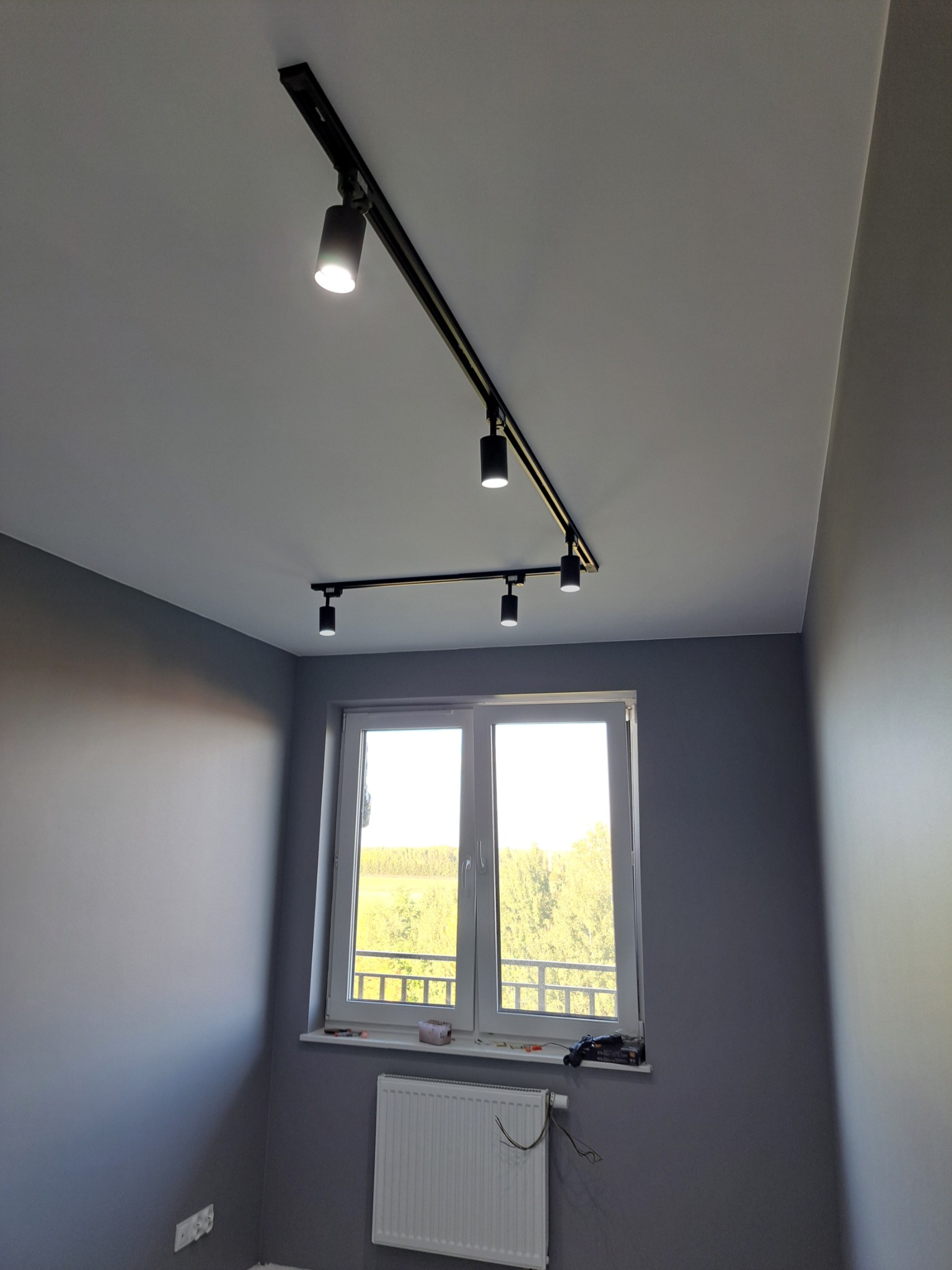 Szare wnętrze pokoju po remoncie z zamontowanym oświetleniem szynowym z reflektorami punktowymi, widok na okno i grzejnik.