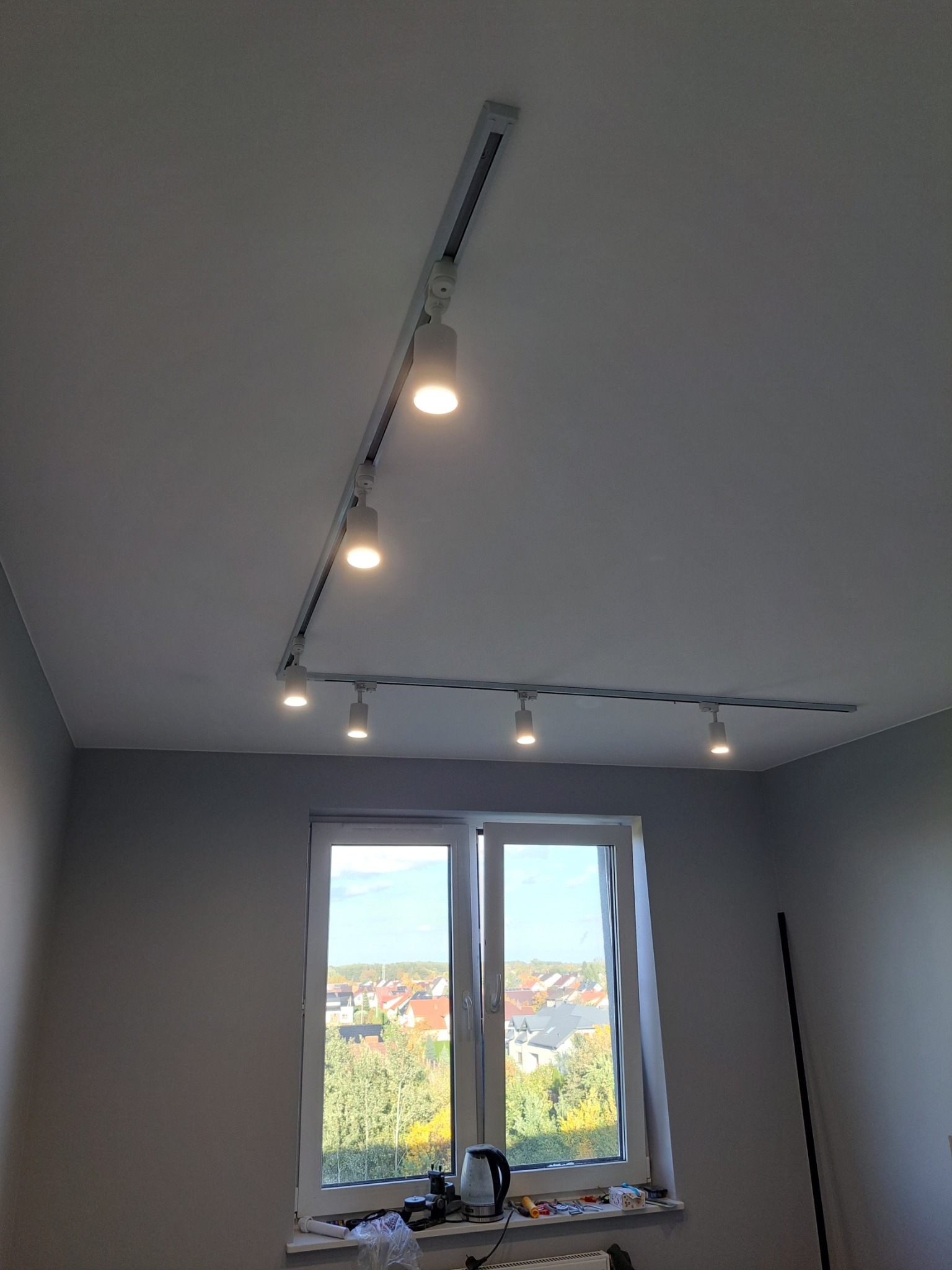 Nowoczesne oświetlenie szynowe z reflektorami punktowymi w szarym pokoju, widok z okna na osiedle domów jednorodzinnych.