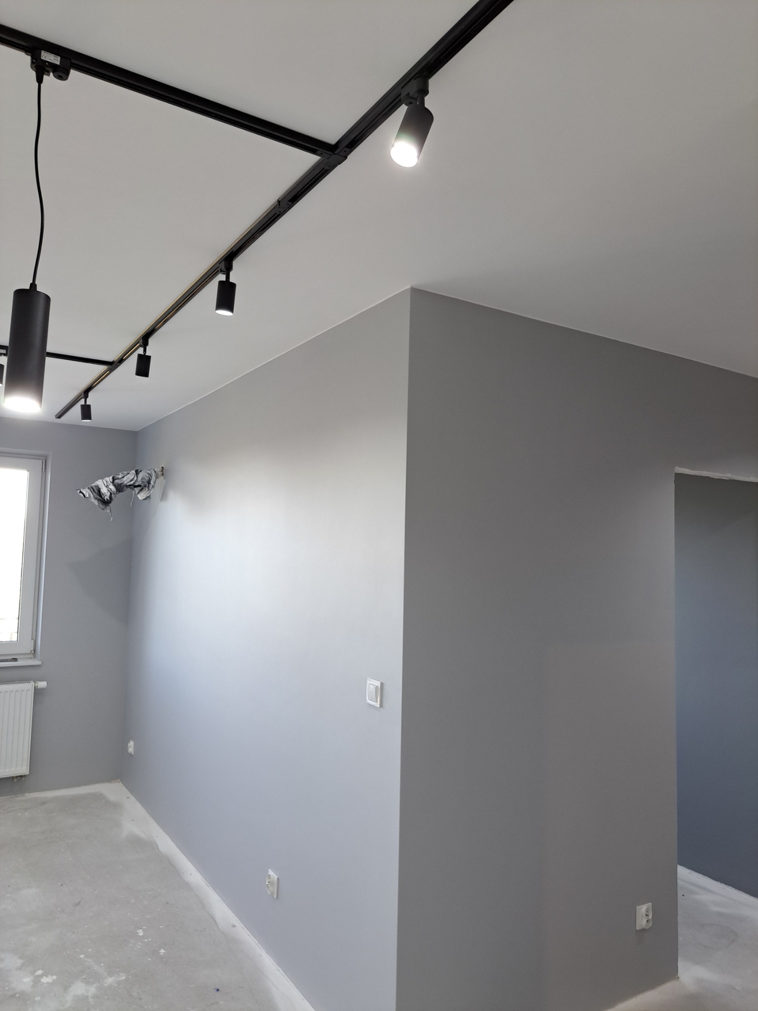 Wnętrze pokoju po malowaniu ścian na szaro, z zamontowanym czarnym systemem szynoprzewodów oświetleniowych na suficie, widoczne gniazdka elektryczne na ścianie i zabezpieczona folią klimatyzacja.