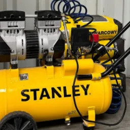 Żółty kompresor Stanley na kółkach, z dwoma cylindrami, na tle worków z zaprawą budowlaną i zwiniętym niebieskim wężem pneumatycznym.