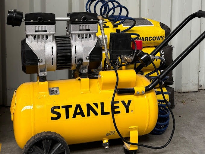 Żółty kompresor Stanley na kółkach, z dwoma cylindrami, na tle worków z zaprawą budowlaną i zwiniętym niebieskim wężem pneumatycznym.