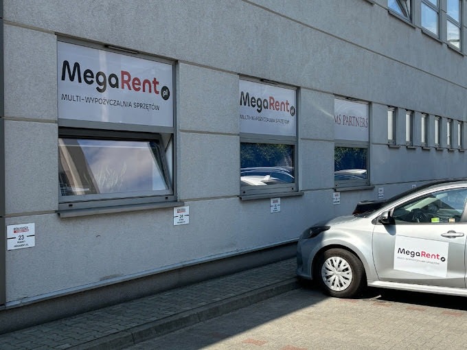 Budynek z oknami z reklamami MegaRent - Multi-Wypożyczalnia Sprzętów i samochód z logo MegaRent na parkingu.