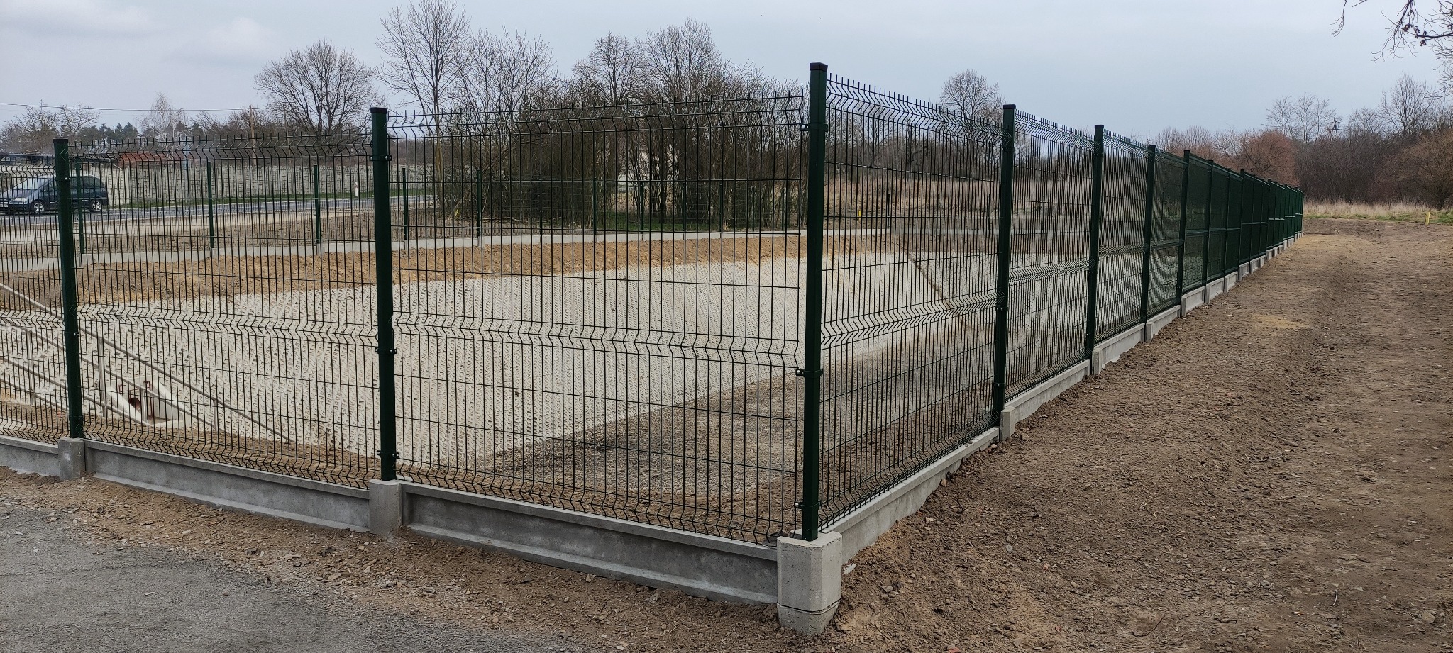 Długi, zielony system ogrodzeniowy z paneli siatkowych, osadzony na betonowej podmurówce, otaczający teren z utwardzoną nawierzchnią i widocznymi drzewami w tle.