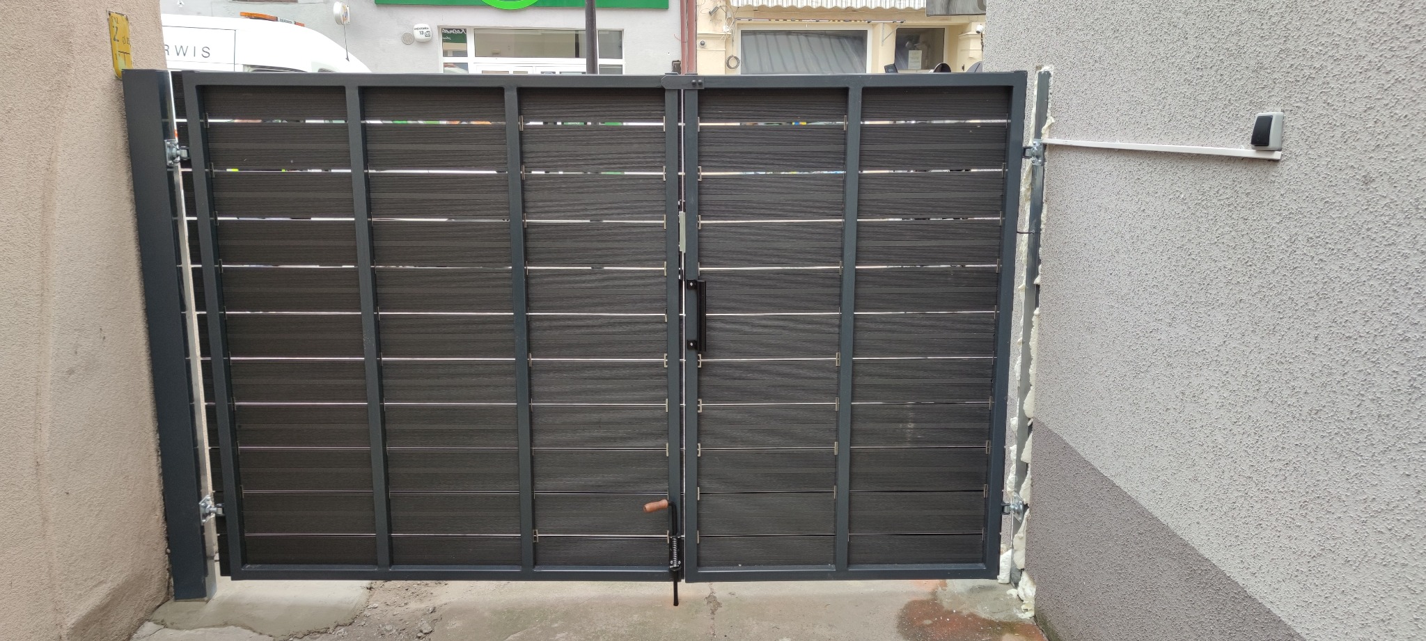 Nowoczesna dwuskrzydłowa brama wjazdowa z poziomymi panelami kompozytowymi w kolorze grafitowym, zamontowana między budynkami, widoczne detale mocowania i pianka montażowa.