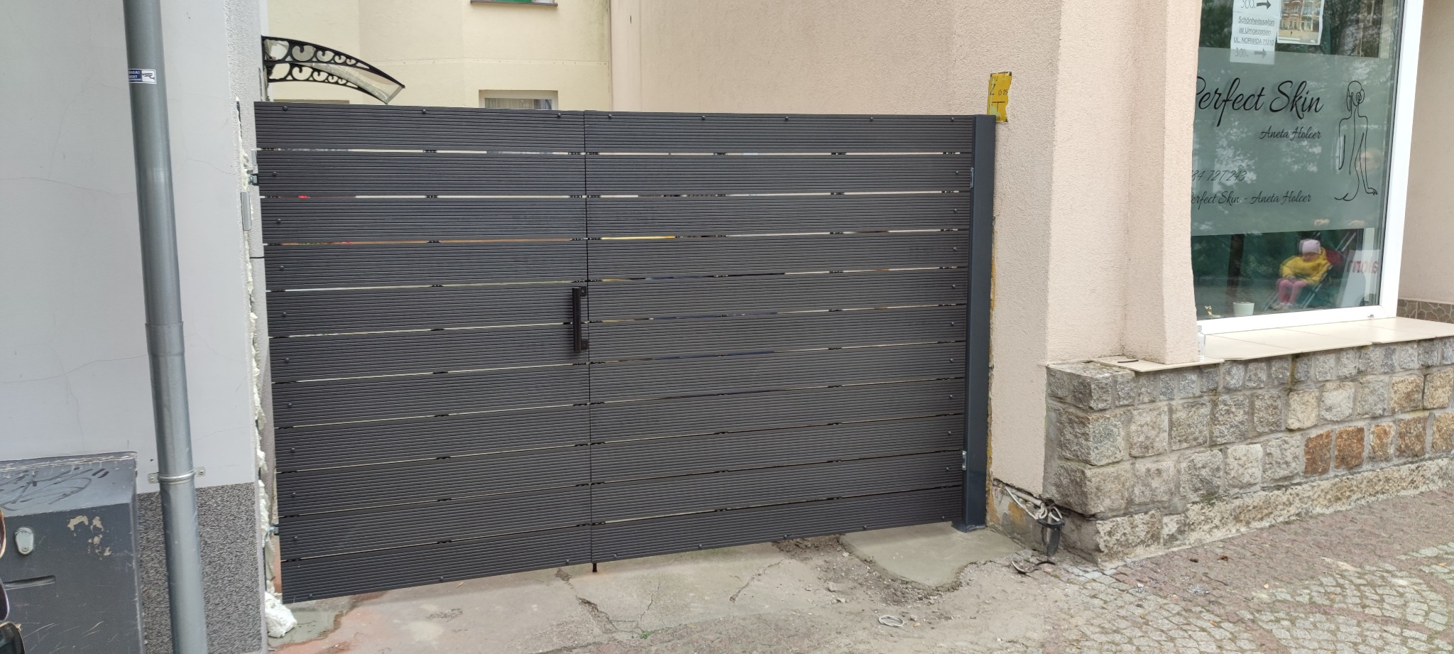 Szara brama wjazdowa z poziomych desek kompozytowych, zamontowana między budynkami, z widoczną klamką i żółtą naklejką na ścianie.