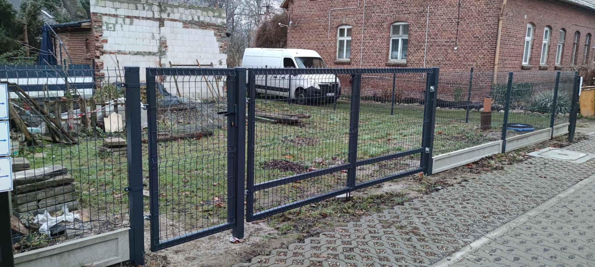 Czarna brama panelowa z uchwytem, otwarta na posesję z widocznym białym busem i ceglanym budynkiem w tle.