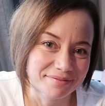 Portret kobiety z krótkimi, brązowymi włosami, lekko uśmiechniętej, z delikatnym makijażem, na rozmytym tle w odcieniach szarości i różu.