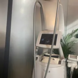 Profesjonalny, biały aparat do endermologii LPG z ekranem dotykowym i metalowymi rurami, widoczne akcesoria i logo LPG, na tle ściany i rośliny doniczkowej.