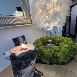 Gabinet Kosmetyczny Monika Tomczuk - Zapachowa świeca w ceramicznej głowie, obok wieniec z mchu i lampa z piór. Na ścianie wisi lustro w srebrnej ramie oraz zdjęcie kobiety.