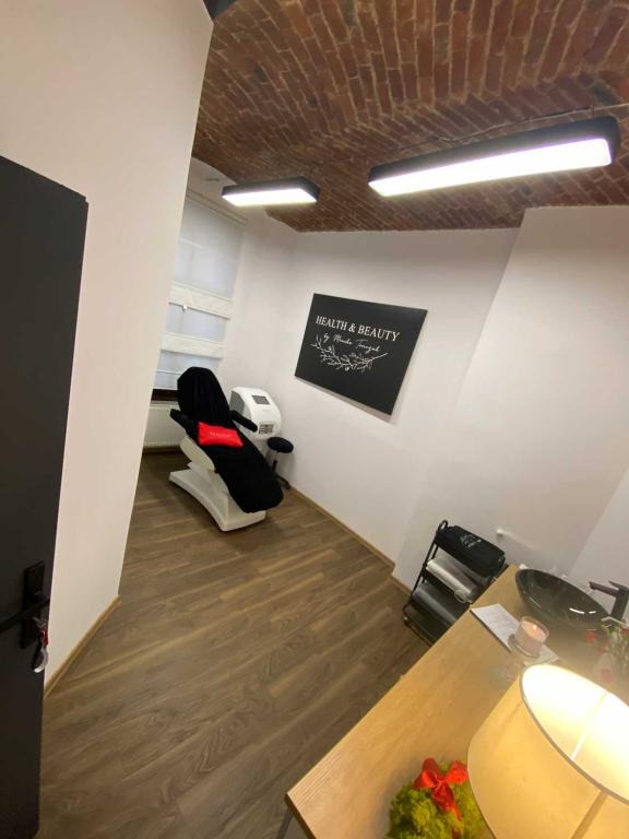 Wnętrze gabinetu kosmetycznego z ceglanym sufitem, białym fotelem zabiegowym z czarnym pokrowcem i czerwoną poduszką, oraz sprzętem kosmetycznym w tle. Na ścianie wisi czarna tablica z napisem...