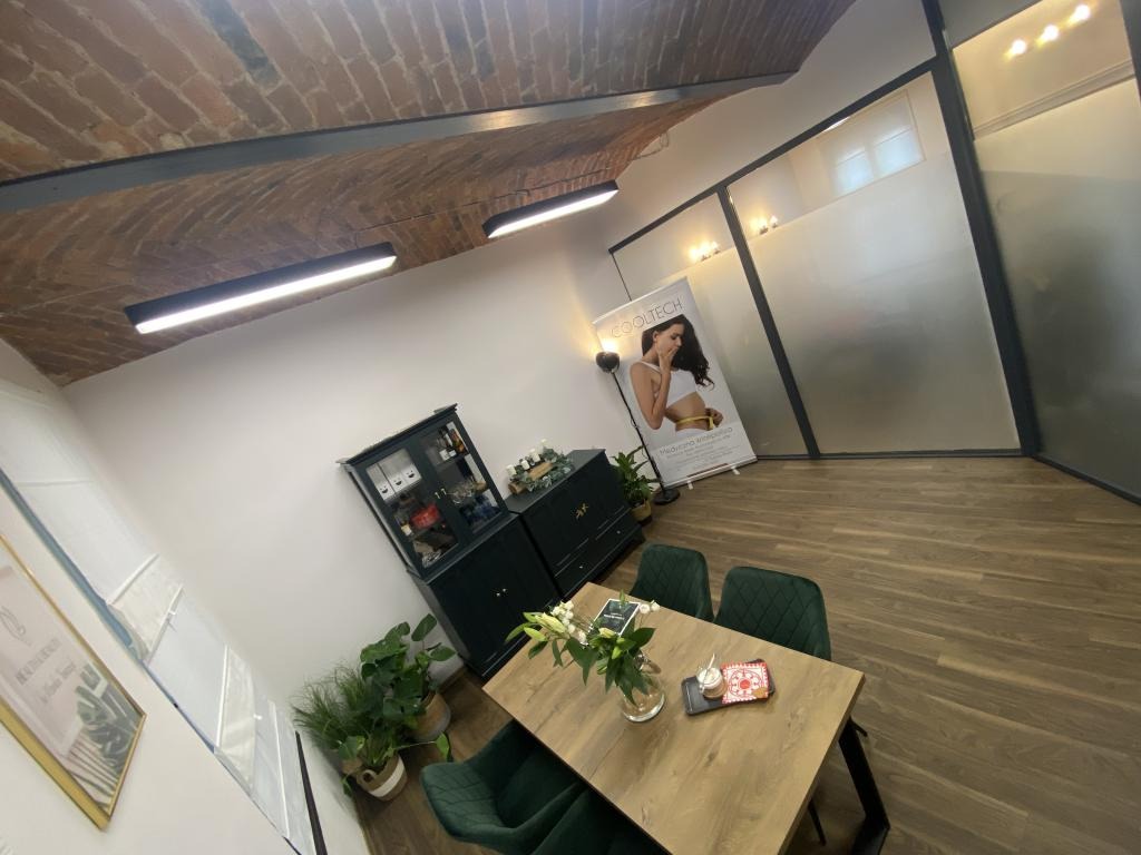 Wnętrze gabinetu kosmetycznego z ceglanym sklepieniem, drewnianym stołem z kwiatami, ciemnozielonymi krzesłami i plakatem reklamowym zabiegów na ciało.