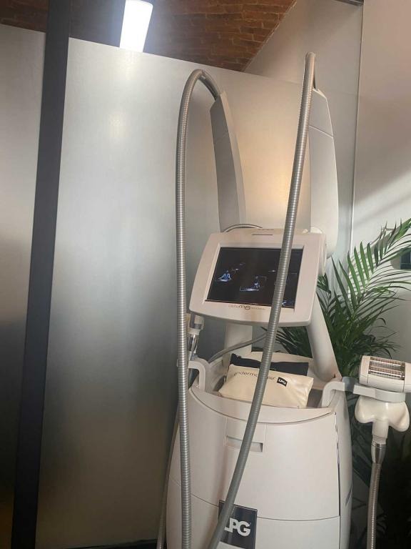 Profesjonalny, biały aparat do endermologii LPG z ekranem dotykowym i metalowymi rurami, widoczne akcesoria i logo LPG, na tle ściany i rośliny doniczkowej.