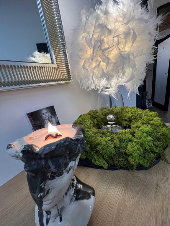 Zapachowa świeca w ceramicznej głowie, obok wieniec z mchu i lampa z piór. Na ścianie wisi lustro w srebrnej ramie oraz zdjęcie kobiety.