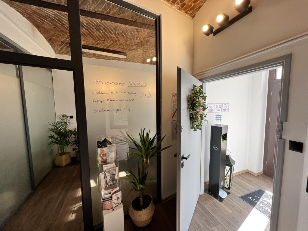 Nowoczesne wnętrze salonu z tablicą informującą o promocjach na zabiegi, widoczne rośliny doniczkowe i dyspenser do dezynfekcji.
