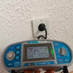 Miernik instalacji elektrycznej Metrel MI 3100 SE podłączony do gniazdka elektrycznego, wyświetlający wynik pomiaru impedancji pętli zwarcia na tle ściany z fakturą.