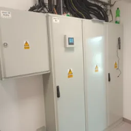 Rozdzielnica elektryczna z licznikiem na ścianie, widoczne kable w korytkach i butelka wody na górze.