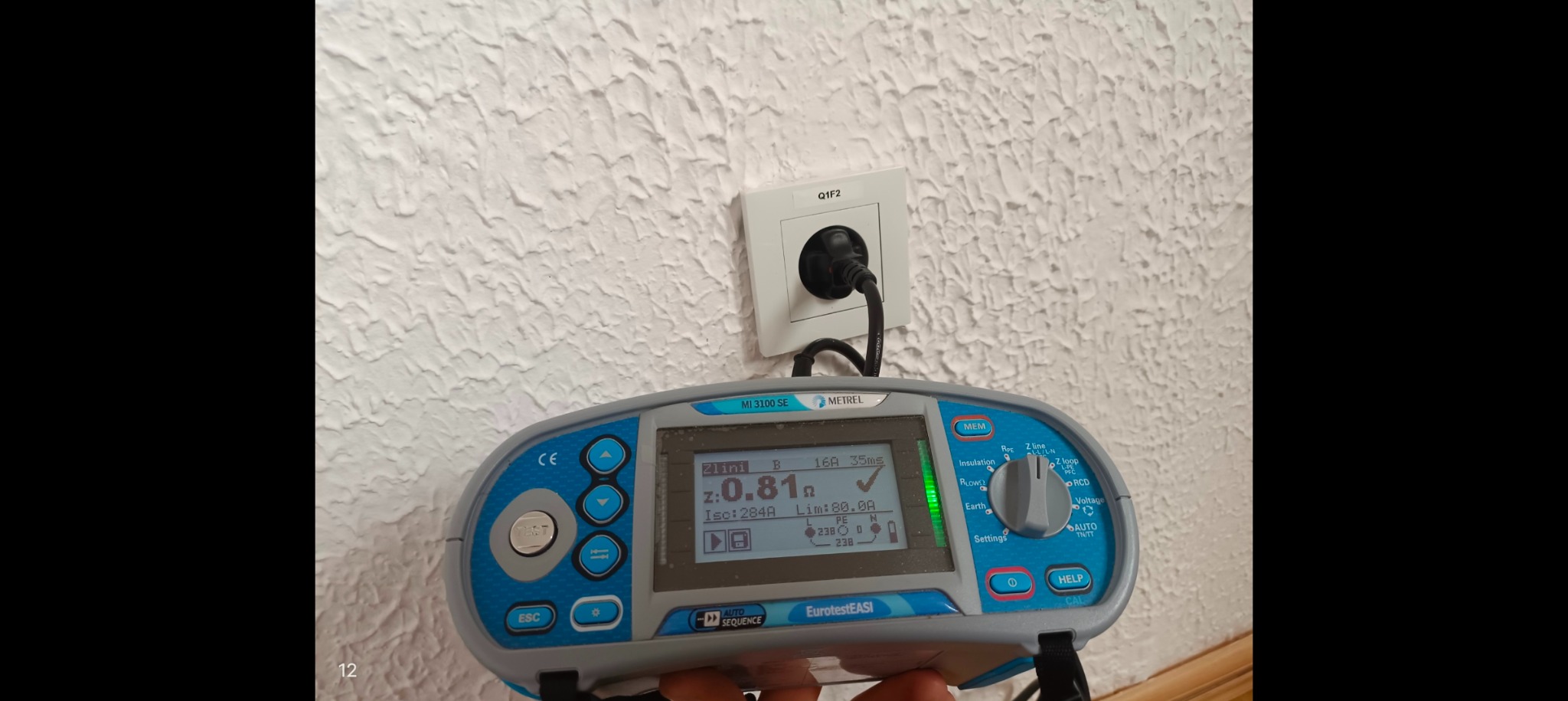 Miernik instalacji elektrycznej Metrel MI 3100 SE podłączony do gniazdka elektrycznego, wyświetlający wynik pomiaru impedancji pętli zwarcia na tle ściany z fakturą.