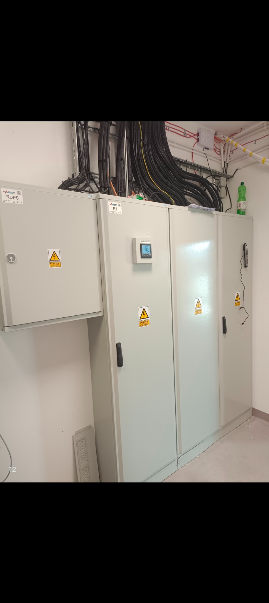 Rozdzielnica elektryczna z licznikiem na ścianie, widoczne kable w korytkach i butelka wody na górze.