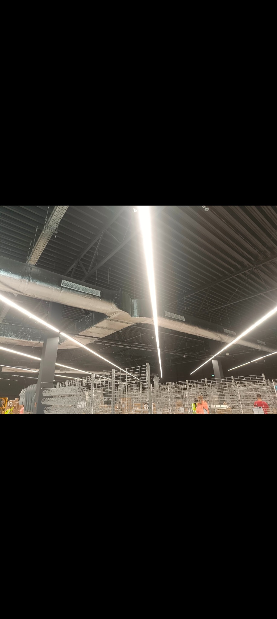 Wnętrze hali magazynowej z widoczną instalacją oświetleniową, kratkami wentylacyjnymi pod sufitem oraz metalowymi regałami i pracownikami w tle.